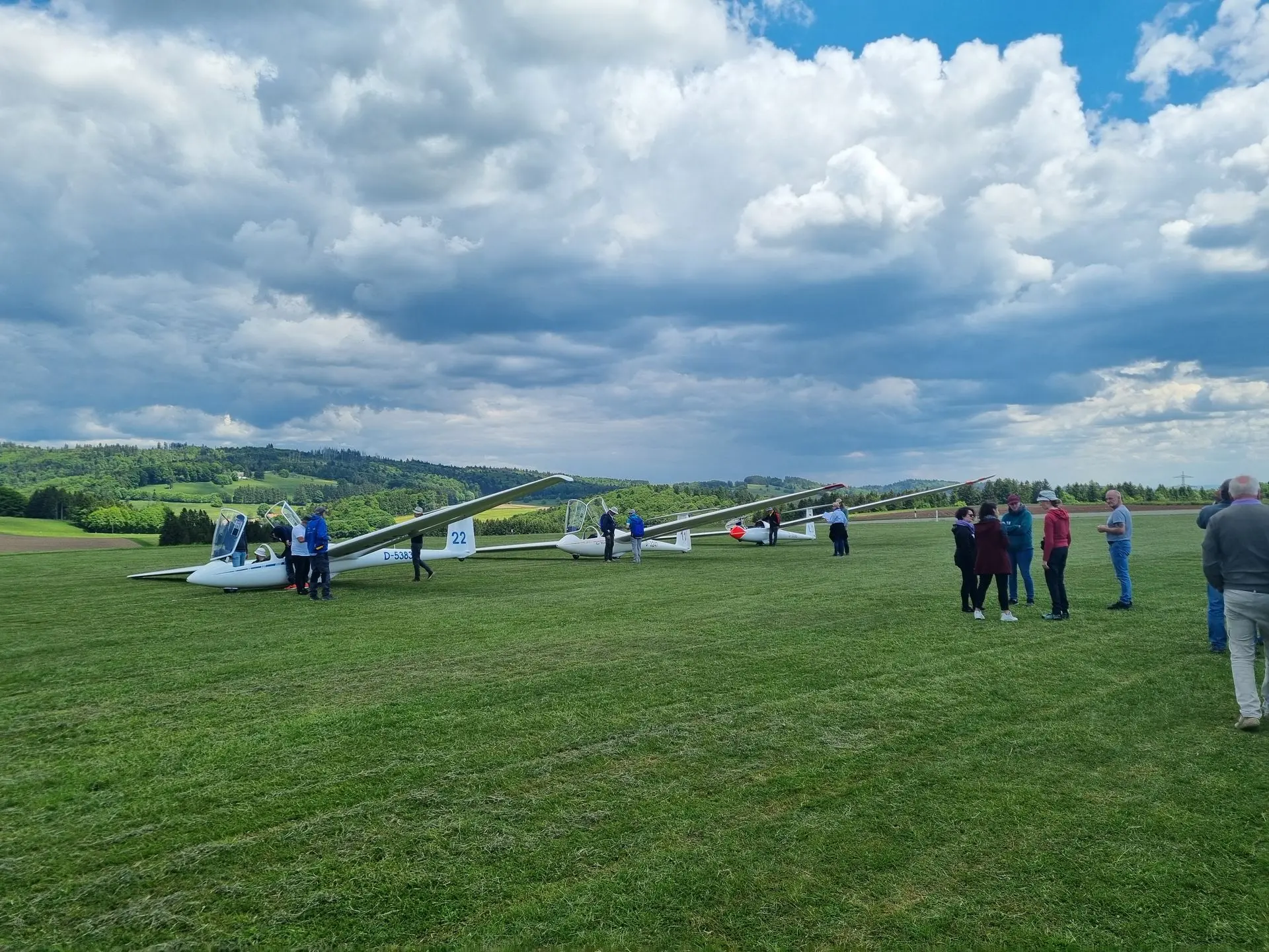 Drei doppelsitzige Segelflugzeuge stehen auf einer Wiese, jeweils Leute am Flugzeug, am Himmel eindrucksvolle Cumulusbewölkung.