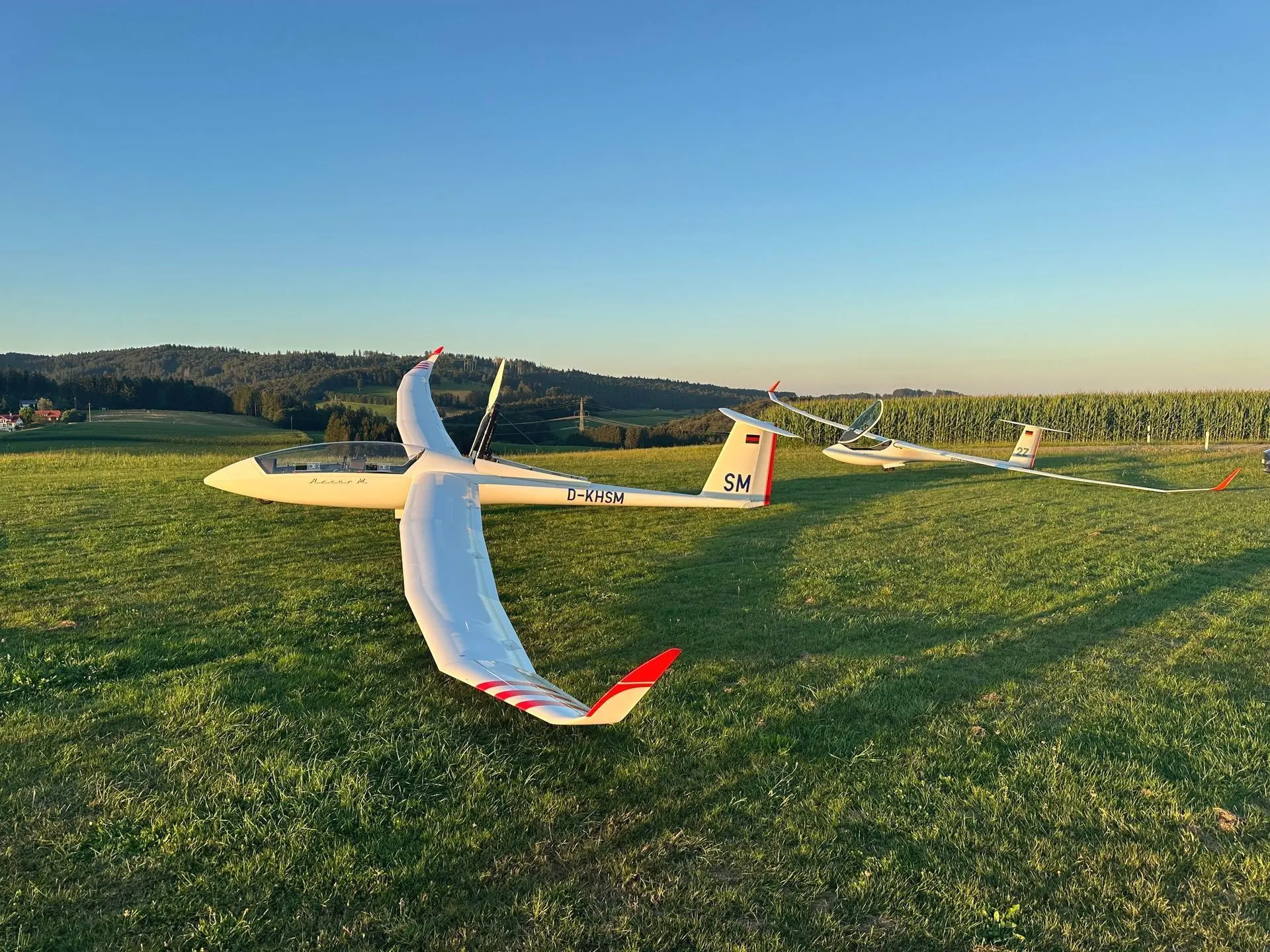 Zwei Segelflugzeuge, jeweils weiß mit roten Tragflächenspitzen, auf einer Wiese. Das vordere ist ein Doppelsitzer und hat den Propeller ausgefahren, das hintere ist ein Einsitzer und hat die Haube offen.