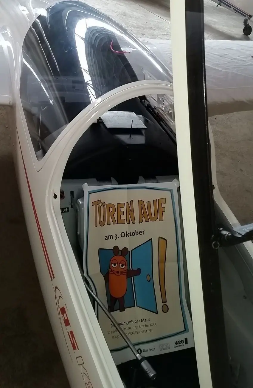 Eine Flugzeughalle. Eine ASK 21 mit geöffneter Haube. Auf dem Sitz liegt ein Plakat der Sendung mit der Maus. Aufschrift: Türen auf am 3. Oktober.