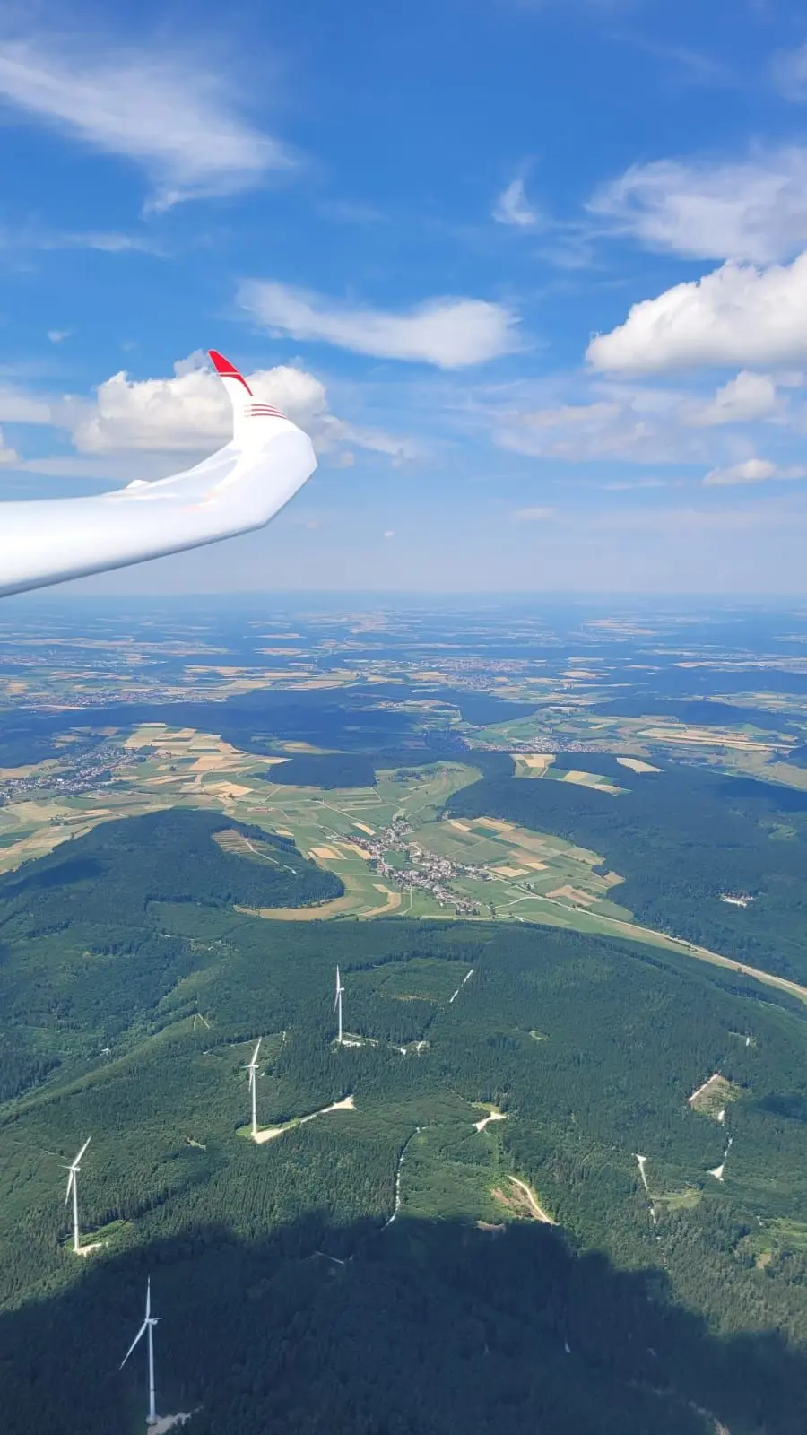 Die Tragfläche eines Segelflugzeugs, darunter Landschaft und Wälder mit Windkraftanlagen.